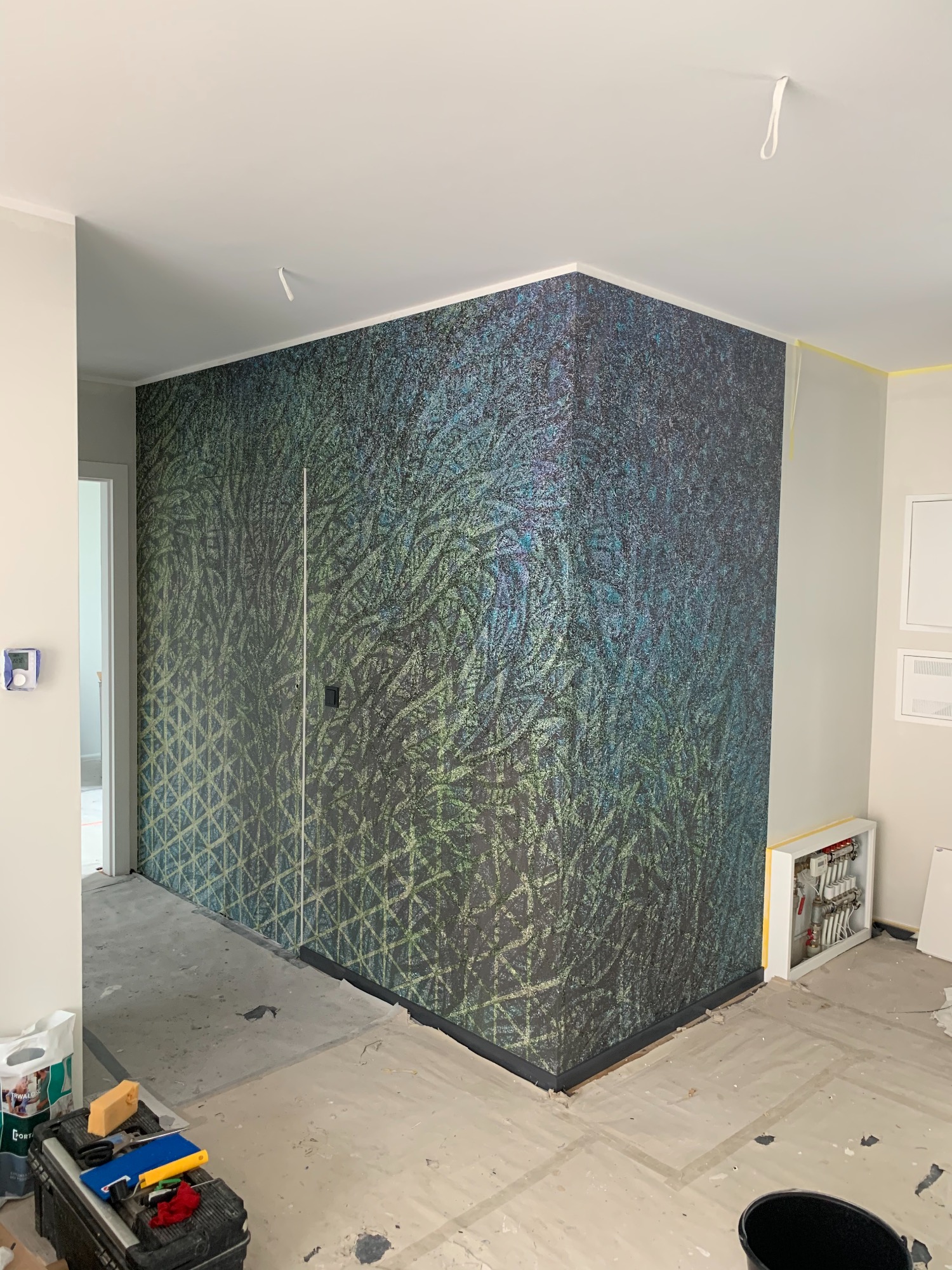 Ściana w pokoju po remoncie z tapetą w odcieniach zieleni i niebieskiego o wzorze przypominającym liście, widoczny fragment instalacji grzewczej w białej obudowie i narzędzia w skrzynce.
