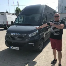 Mężczyzna w okularach słonecznych i koszulce z nadrukiem wskazuje na szary samochód dostawczy marki Iveco zaparkowany na brukowanym placu, w tle inne busy i budynek.
