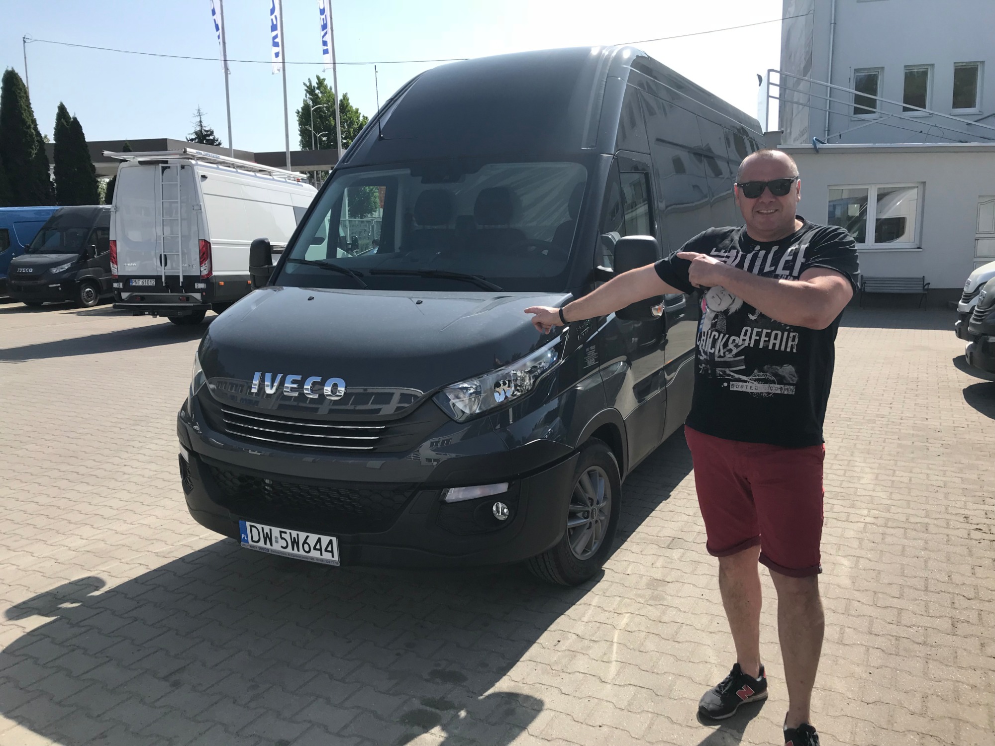 Mężczyzna w okularach słonecznych i koszulce z nadrukiem wskazuje na szary samochód dostawczy marki Iveco zaparkowany na brukowanym placu, w tle inne busy i budynek.