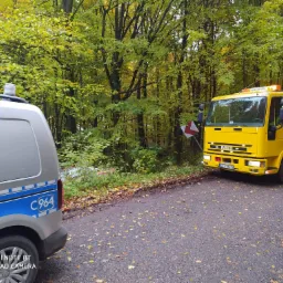 Żółta laweta Iveco z włączonymi światłami awaryjnymi stoi na poboczu leśnej drogi, obok srebrnego samochodu pomocy drogowej z niebieskim pasem i napisem C964.