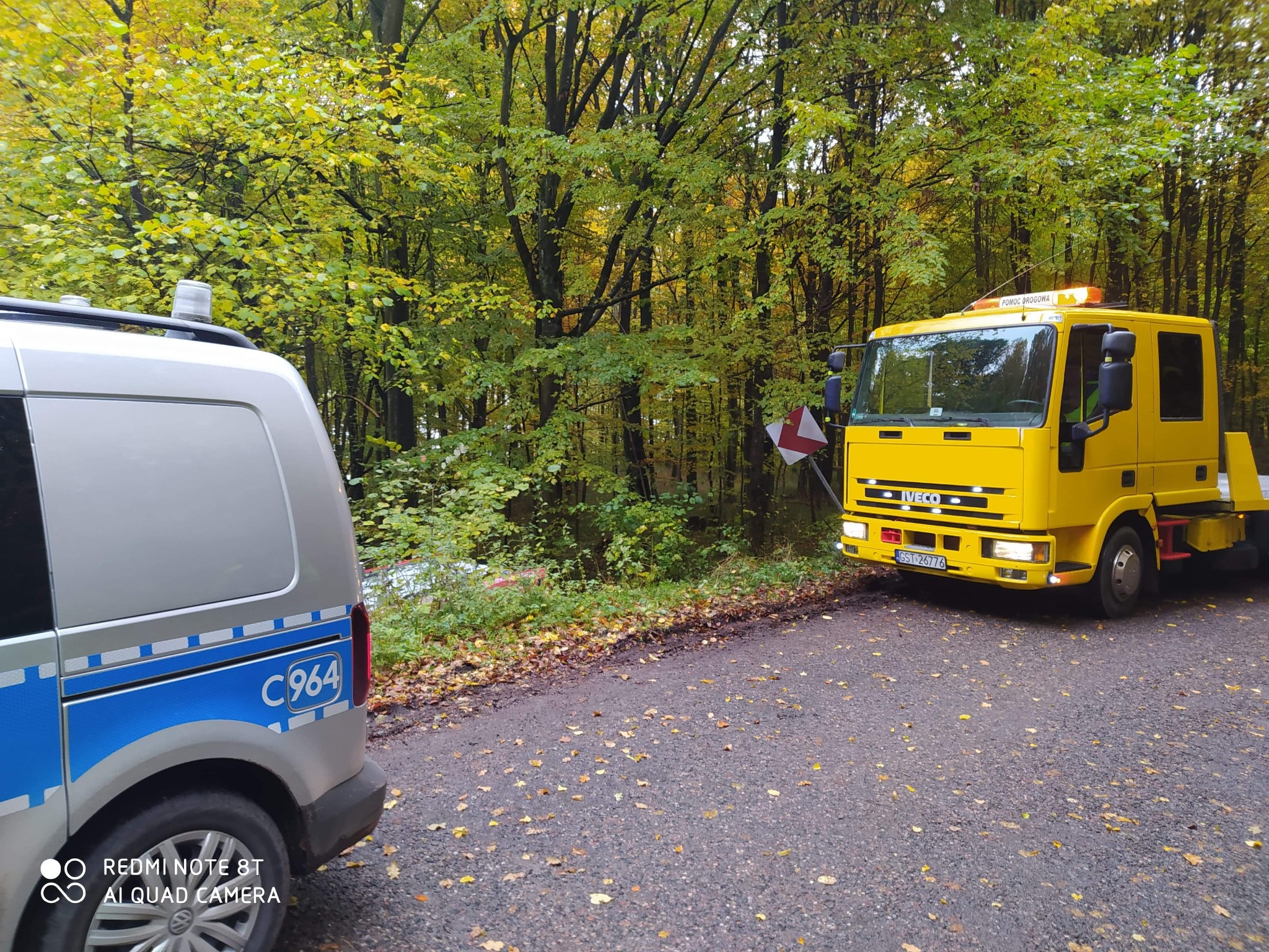 Żółta laweta Iveco z włączonymi światłami awaryjnymi stoi na poboczu leśnej drogi, obok srebrnego samochodu pomocy drogowej z niebieskim pasem i napisem C964.