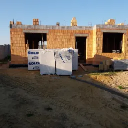 Surowy stan budynku z czerwonej cegły z otworami okiennymi i drzwiowymi, widoczne materiały izolacyjne SOLBET, ślady opon na piaszczystym podłożu.