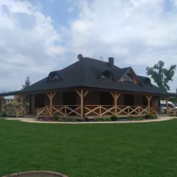 Gotowy, parterowy budynek z drewna z czarnym dachem, otoczony równo przystrzyżonym trawnikiem i niskimi rabatami kwiatowymi, widok z przodu.