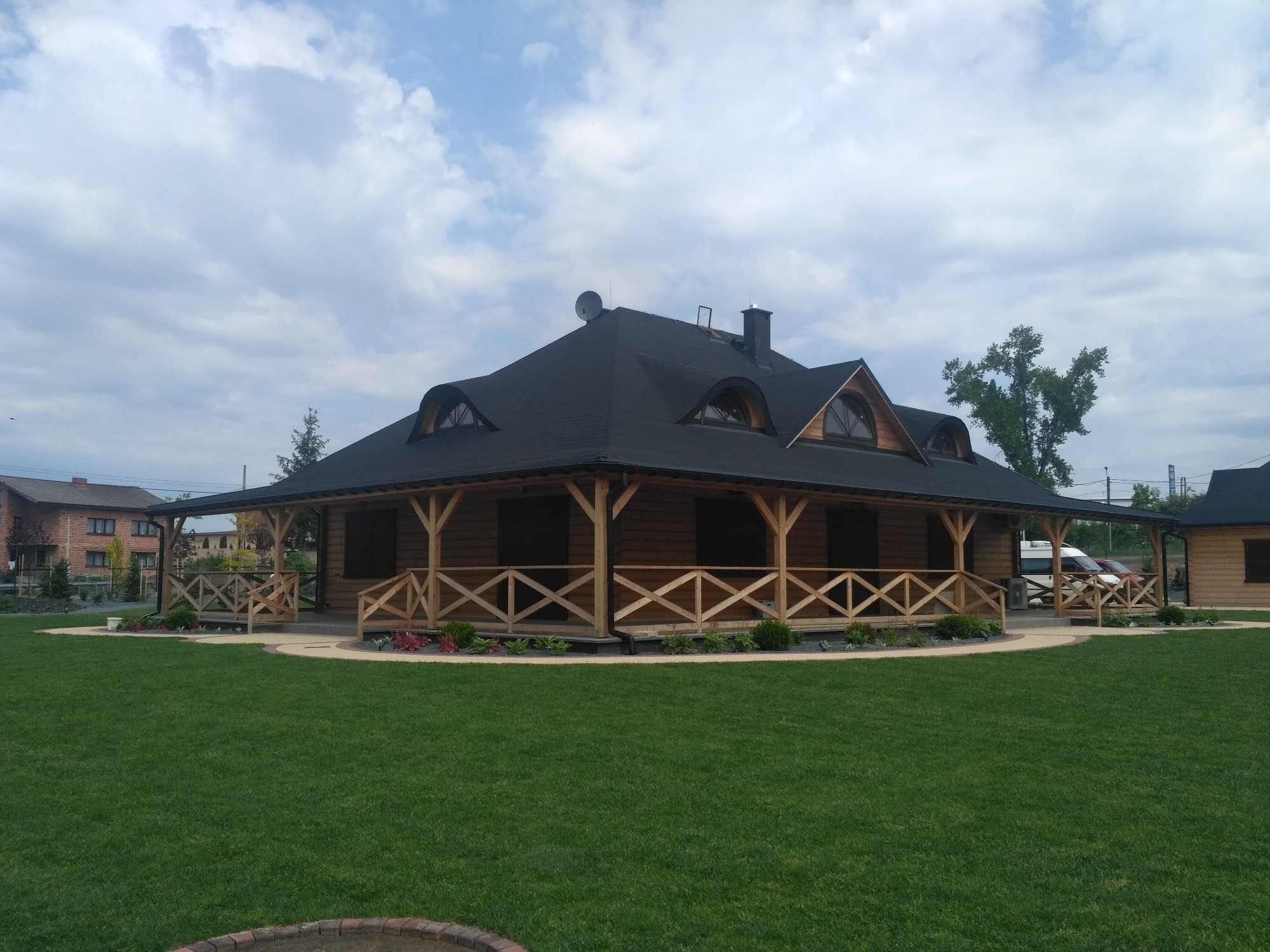 Gotowy, parterowy budynek z drewna z czarnym dachem, otoczony równo przystrzyżonym trawnikiem i niskimi rabatami kwiatowymi, widok z przodu.