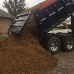 Wyładunek brązowej ziemi z wywrotki na tle domów jednorodzinnych.