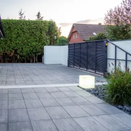 Nowoczesny podjazd z szarych płyt betonowych, oświetlony kostką LED przy ozdobnym nasadzeniu trawy, z bramą w tle i białym murem.