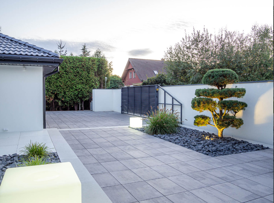 Nowoczesny podjazd z szarej kostki brukowej, oświetlony lampami LED, z dekoracyjnym drzewkiem bonsai i czarnym grysem, brama w tle.