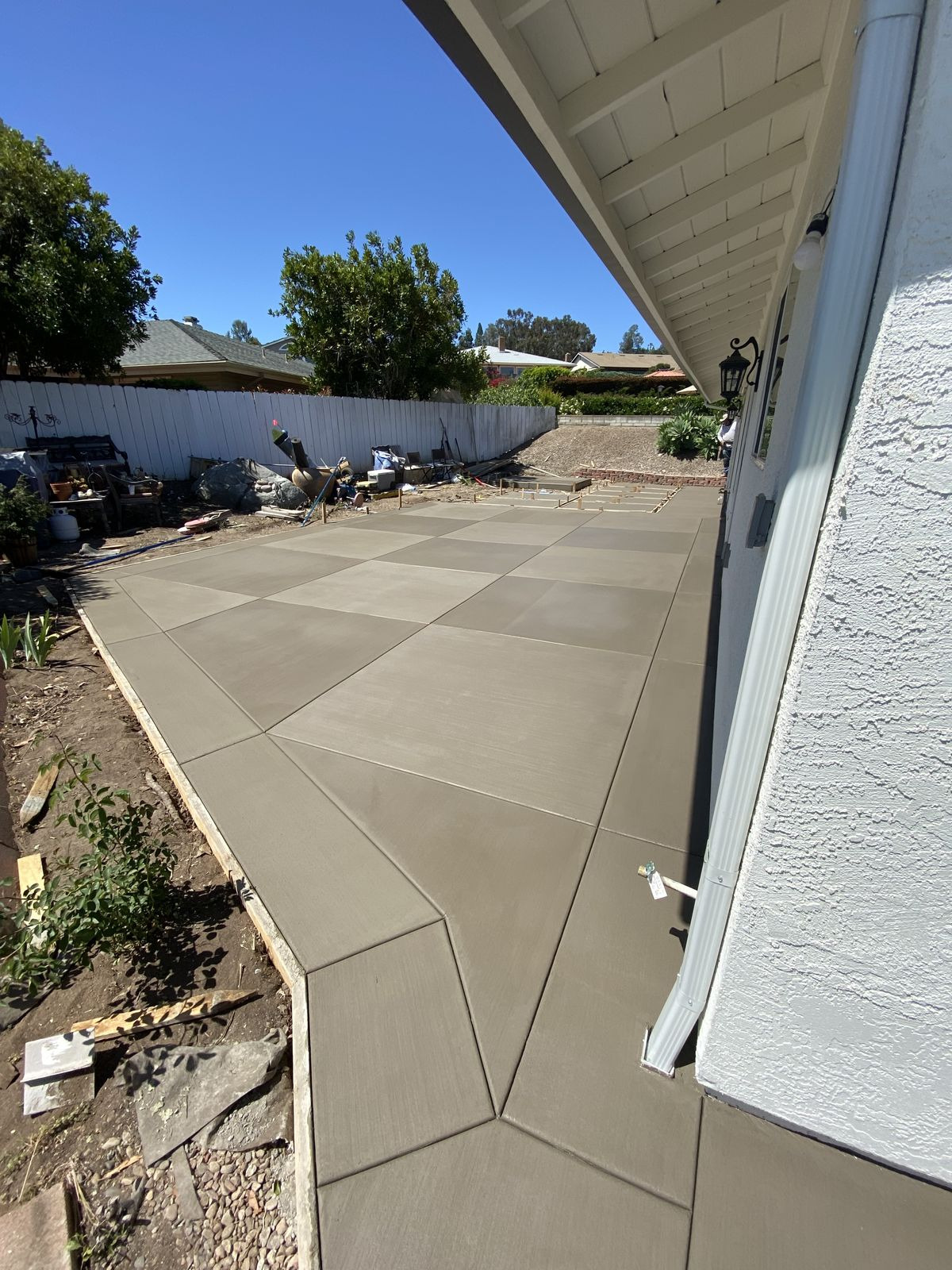 Świeżo wylana, gładka betonowa posadzka podzielona na kwadratowe sekcje z widocznymi liniami podziału, otoczona drewnianym szalunkiem, z białym ogrodzeniem w tle i fragmentem elewacji budynku po...