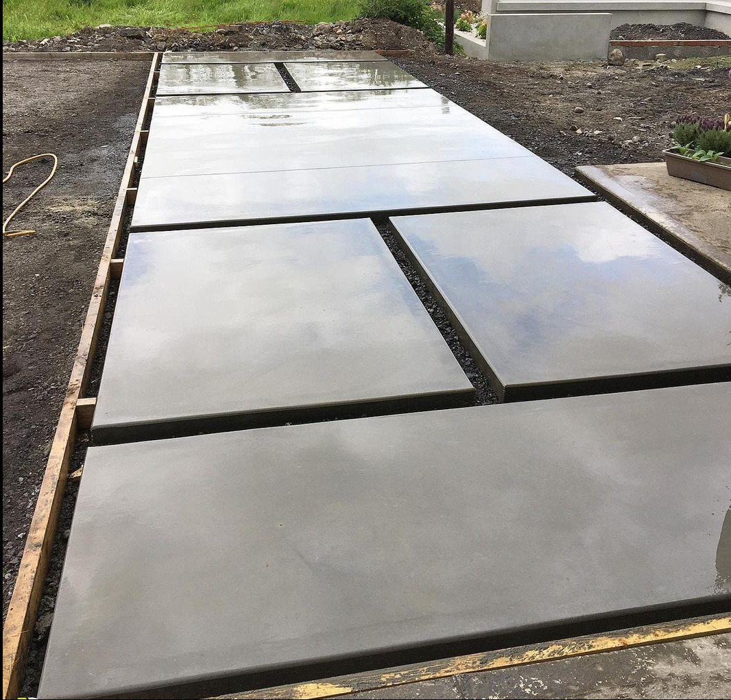 Świeżo wylane, mokre betonowe płyty chodnikowe w drewnianej ramie, z widocznymi odbiciami nieba, oddzielone od siebie wąskimi pasami żwiru.