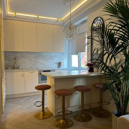 Ewelina Smażyk - Elegancka kuchnia w jasnych barwach z wyspą i hokerami na złotych nogach. Uwagę przykuwa kryształowy żyrandol i dekoracyjny sufit. Całość dopełnia duża roślina doniczkowa.