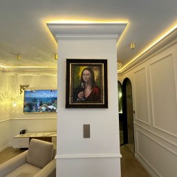 Ewelina Smażyk - Nowoczesne wnętrze z obrazem Mona Lisy z kieliszkiem wina, oświetleniem LED i dekoracyjnymi panelami ściennymi. Widoczny fragment fotela i obraz na ścianie.