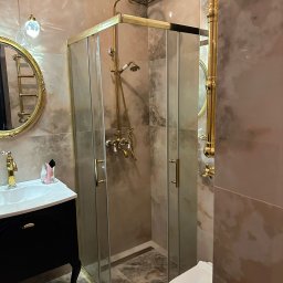 Ewelina Smażyk - Elegancka łazienka z kabiną prysznicową w stylu retro, złote detale, marmurowe płytki i czarna szafka umywalkowa na ozdobnych nóżkach. Lampa wisząca z kryształkami.