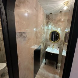 Ewelina Smażyk - Elegancka łazienka z beżowymi płytkami, czarną szafką pod umywalką i okrągłym lustrem w złotej ramie. Wiszące lampy z kryształkami dodają blasku.