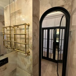 Ewelina Smażyk - Elegancka łazienka z marmurowymi płytkami, złotym grzejnikiem drabinkowym i czarnymi, przeszklonymi drzwiami łukowymi, odbijającymi wnętrze w lustrze.