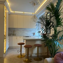 Ewelina Smażyk - Elegancka kuchnia w jasnych barwach z marmurowym blatem, oświetleniem LED i złotymi akcentami. Wysokie krzesła barowe i duża roślina doniczkowa dodają wnętrzu charakteru.
