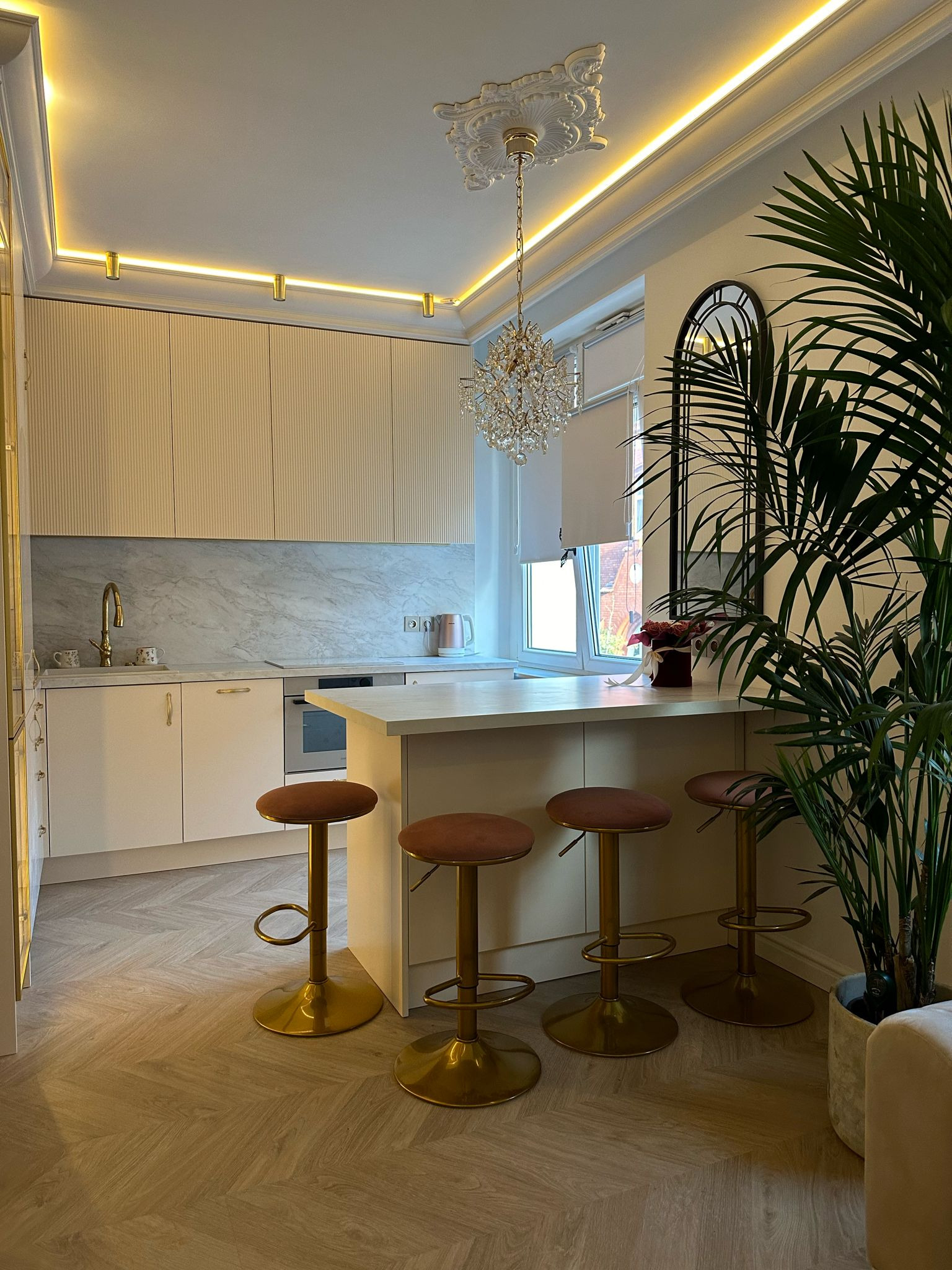 Elegancka kuchnia w jasnych barwach z wyspą i hokerami na złotych nogach. Uwagę przykuwa kryształowy żyrandol i dekoracyjny sufit. Całość dopełnia duża roślina doniczkowa.