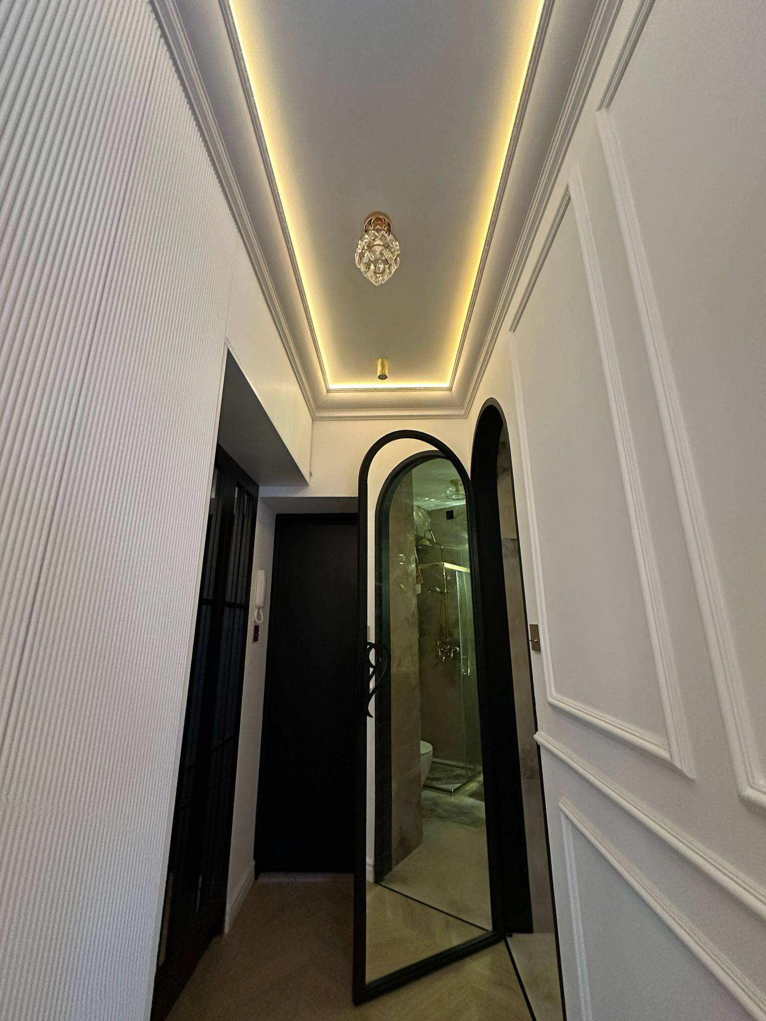 Nowoczesny korytarz z dekoracyjną ścianą, oświetleniem LED i lustrzanymi drzwiami do łazienki. Widoczny fragment łazienki z prysznicem i toaletą.