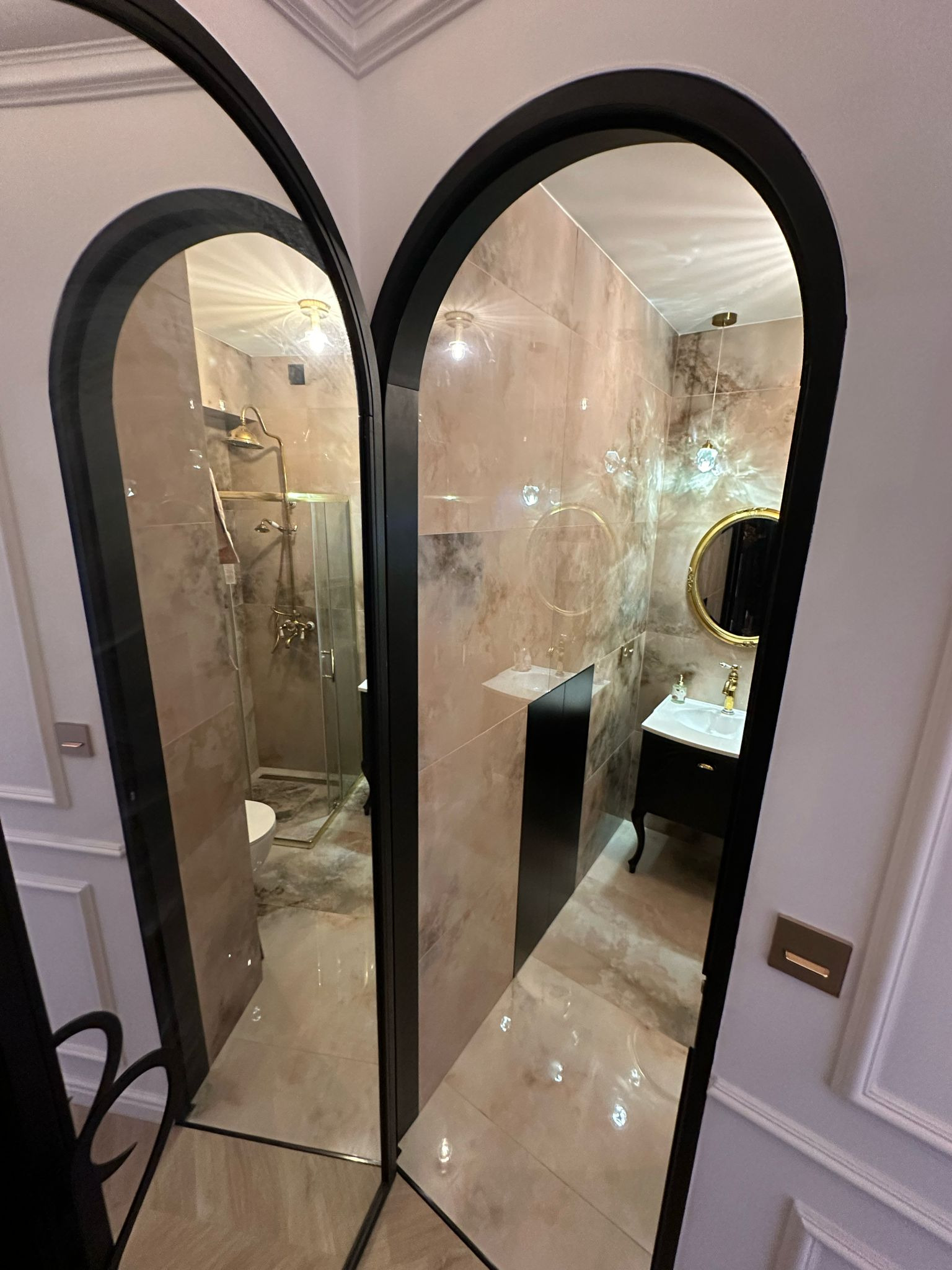 Elegancka łazienka w beżach, widziana przez lustrzane drzwi z czarną ramą. Kabina prysznicowa i umywalka z lustrem w złotych akcentach.
