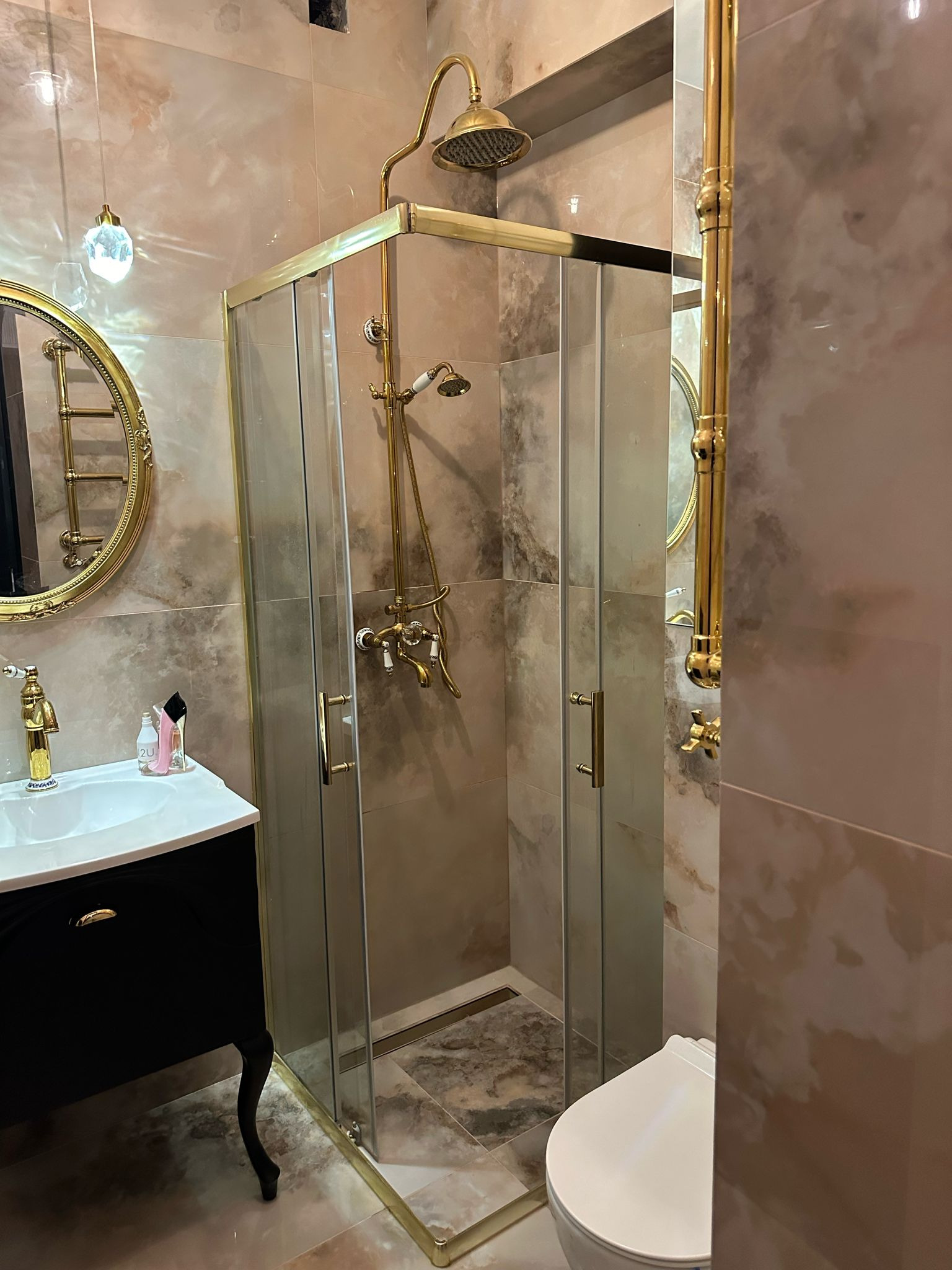 Elegancka łazienka z kabiną prysznicową w stylu retro, złote detale, marmurowe płytki i czarna szafka umywalkowa na ozdobnych nóżkach. Lampa wisząca z kryształkami.