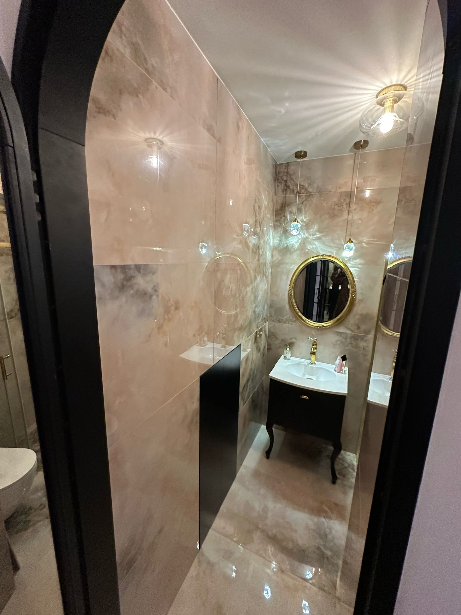 Elegancka łazienka z beżowymi płytkami, czarną szafką pod umywalką i okrągłym lustrem w złotej ramie. Wiszące lampy z kryształkami dodają blasku.