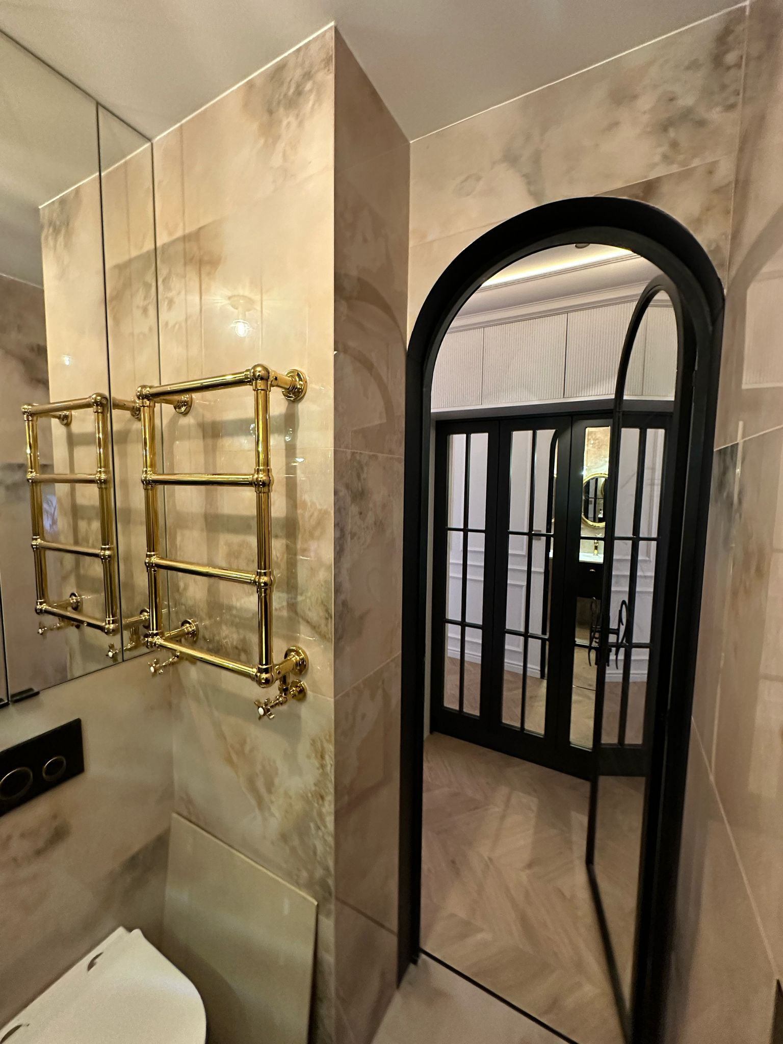 Elegancka łazienka z marmurowymi płytkami, złotym grzejnikiem drabinkowym i czarnymi, przeszklonymi drzwiami łukowymi, odbijającymi wnętrze w lustrze.