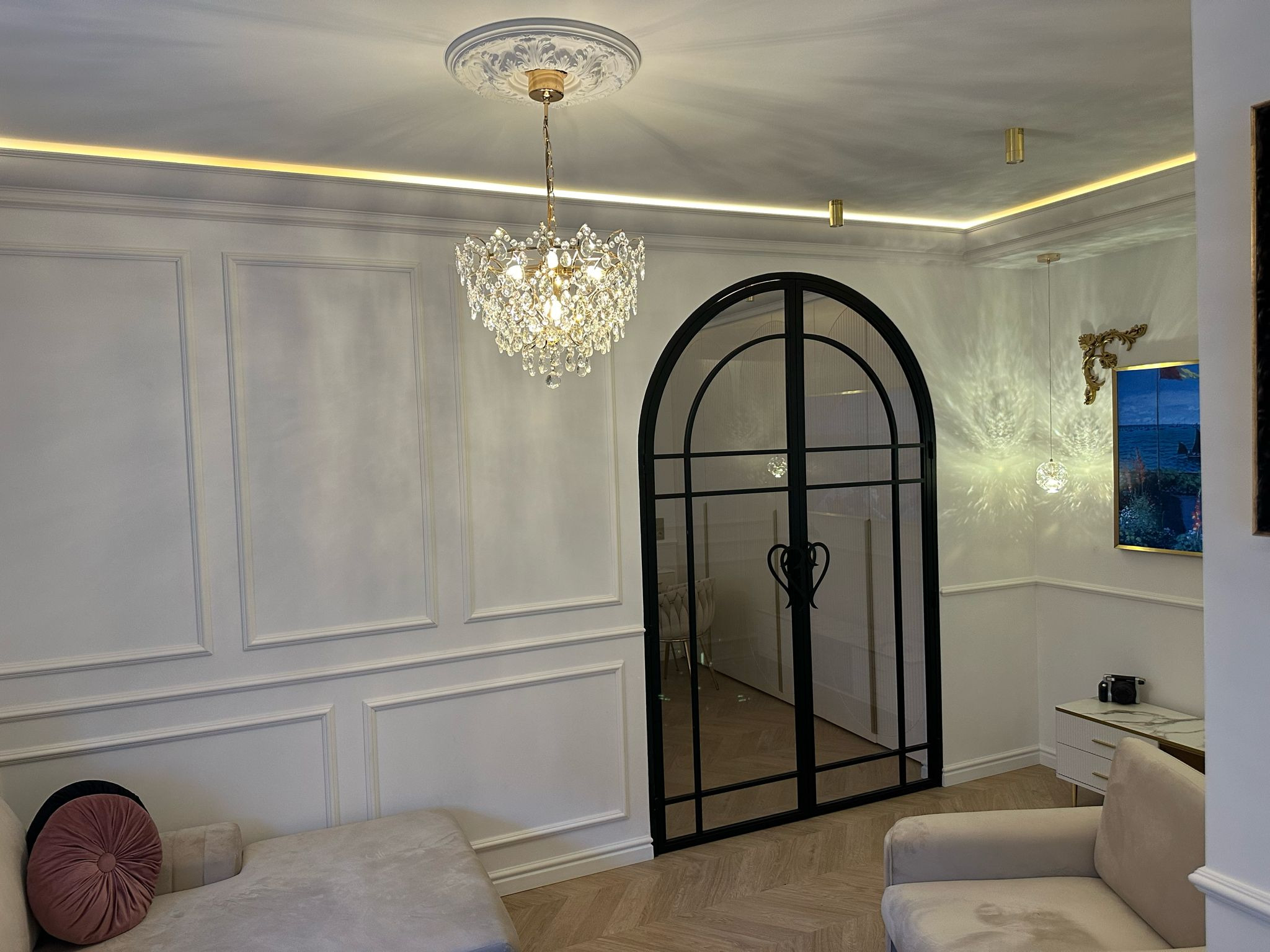 Eleganckie wnętrze: kryształowy żyrandol nad sofą, dekoracyjne listwy ścienne, czarne, łukowe drzwi z serduszkami, parkiet w jodełkę i subtelne oświetlenie LED.