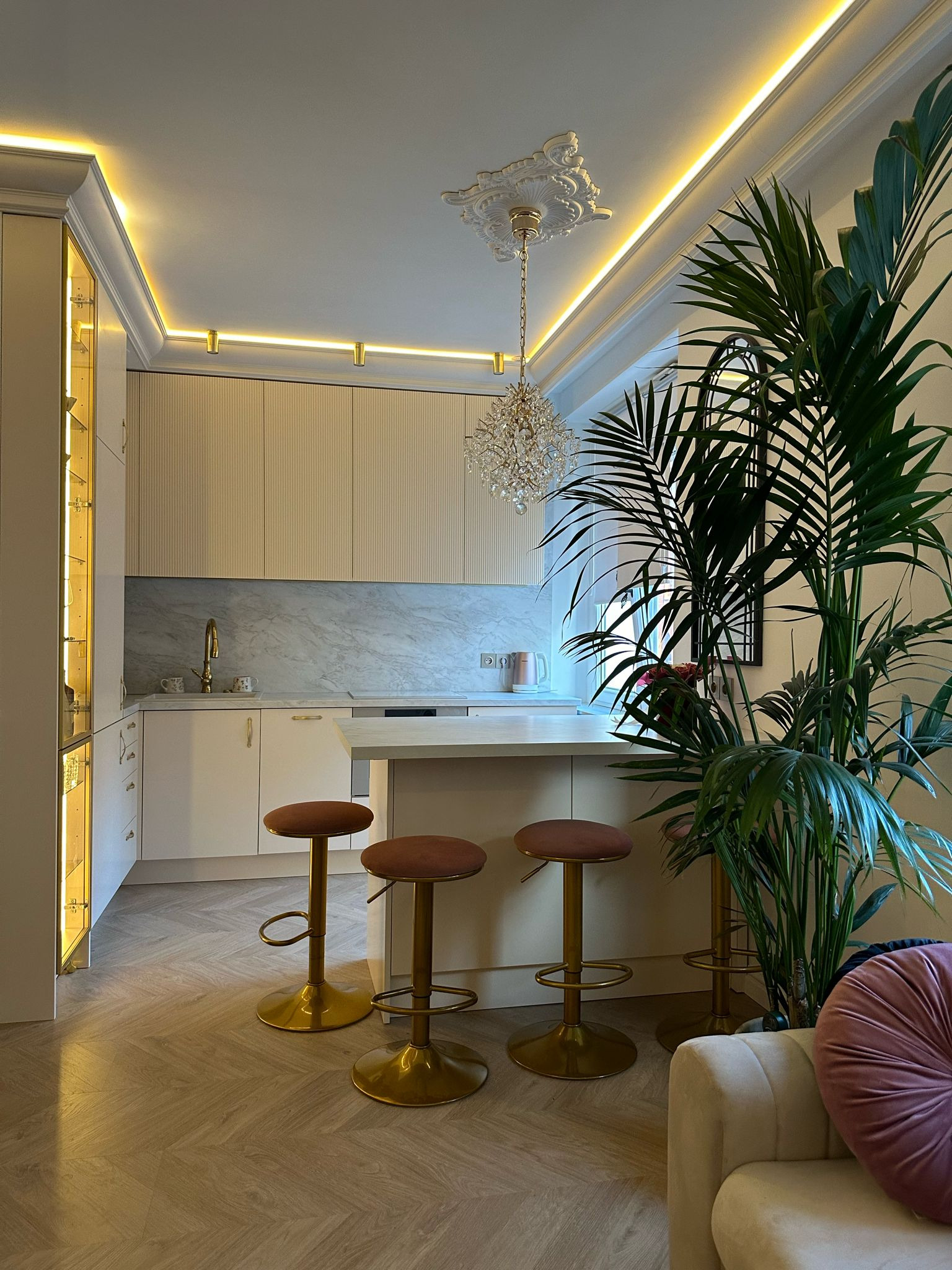 Elegancka kuchnia w jasnych barwach z marmurowym blatem, oświetleniem LED i złotymi akcentami. Wysokie krzesła barowe i duża roślina doniczkowa dodają wnętrzu charakteru.