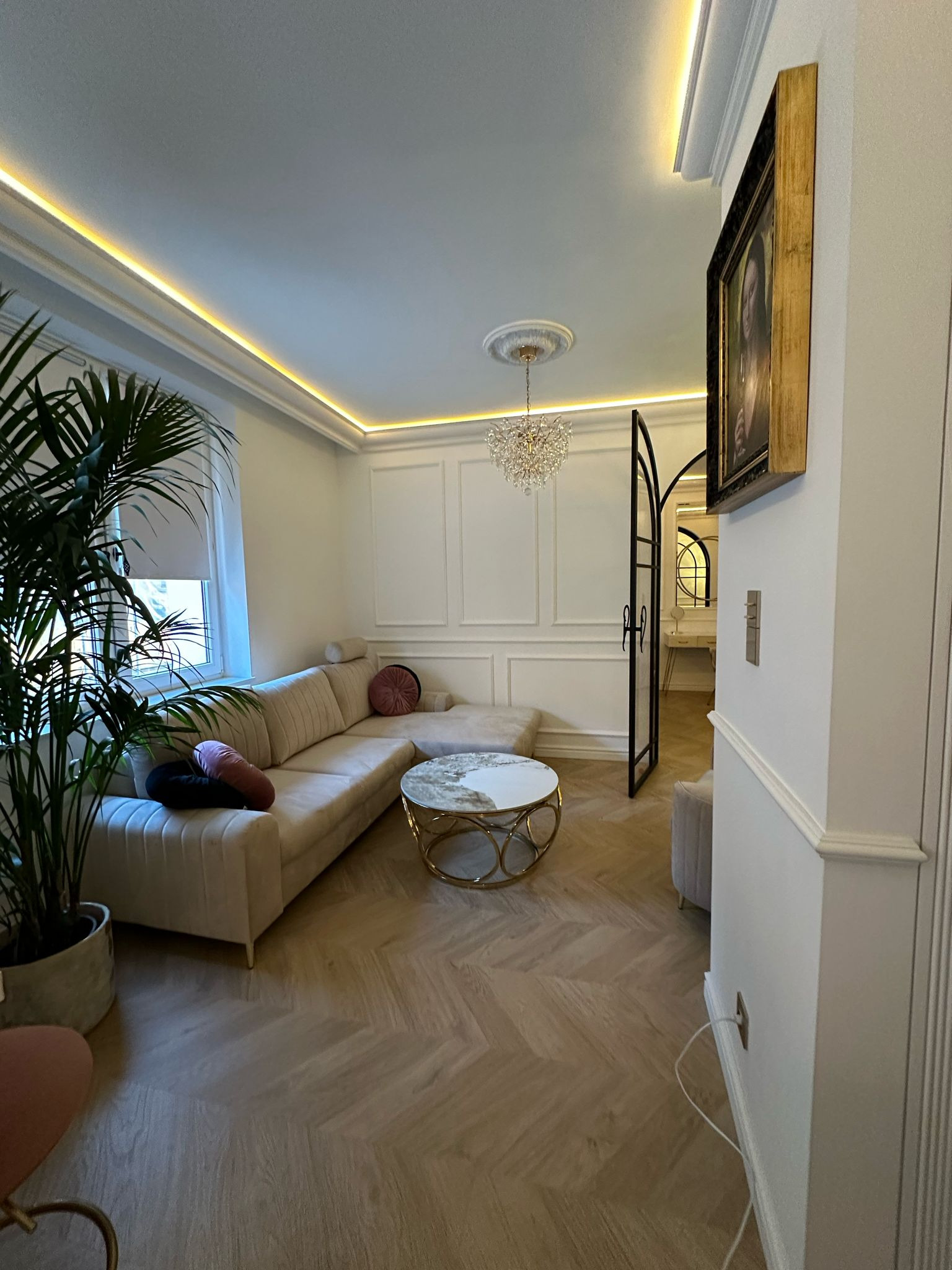 Elegancki salon z narożnikiem w odcieniach beżu, okrągłym stolikiem kawowym ze złotymi akcentami i parkietem w jodełkę. Dekoracyjne listwy ścienne i kryształowy żyrandol.