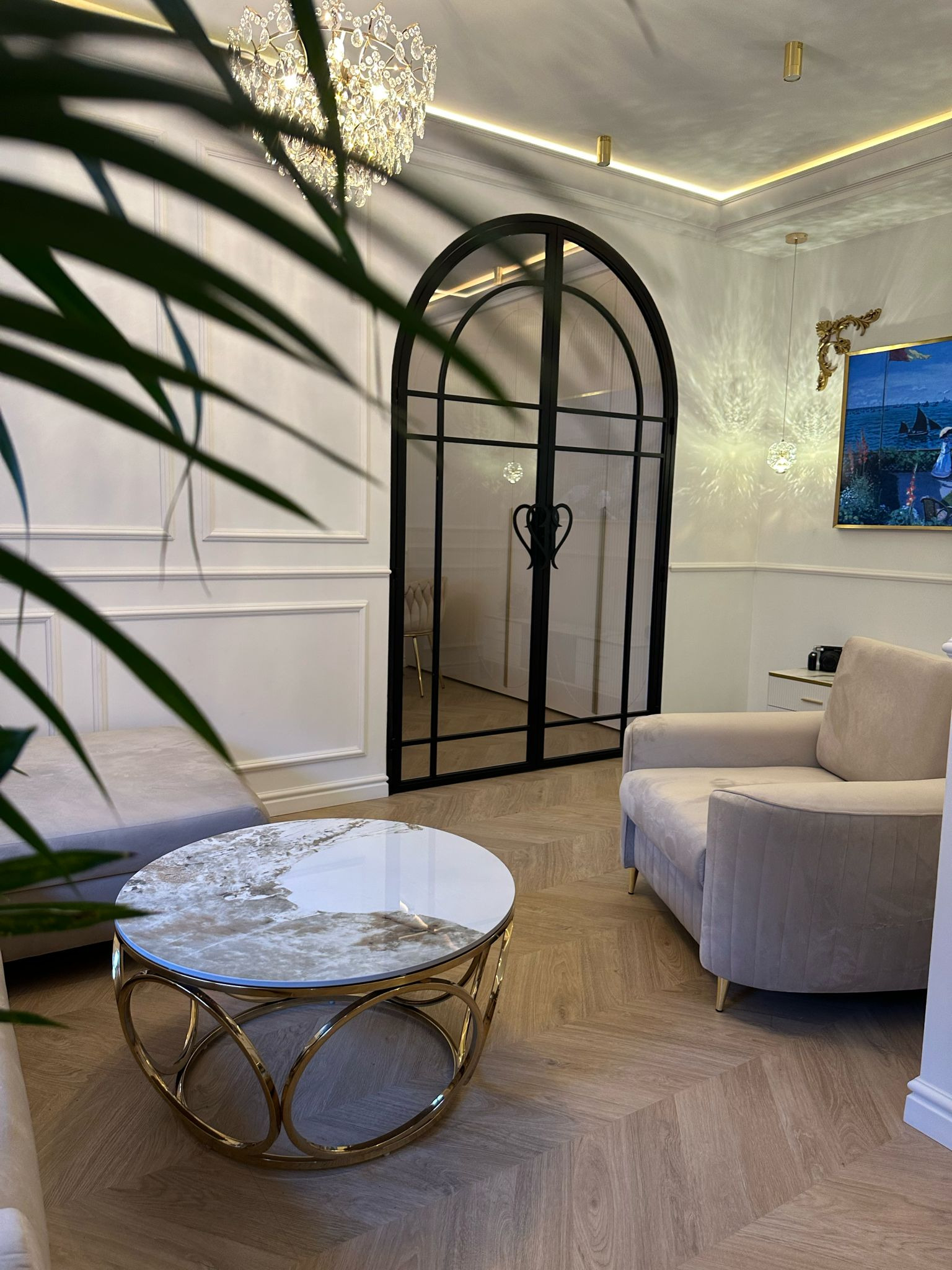 Elegancki salon z parkietem w jodełkę, kremowym fotelem i stolikiem kawowym ze złotą podstawą. W tle czarna, przeszklona ścianka działowa i kryształowy żyrandol.