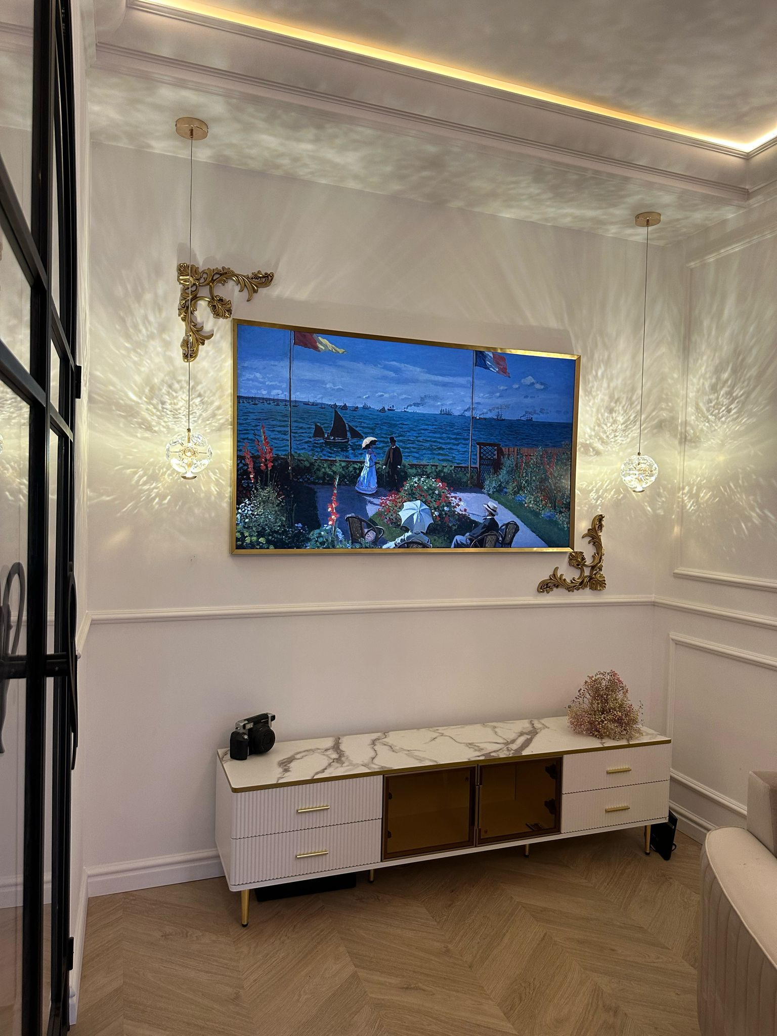 Elegancki salon z obrazem w złotej ramie nad białą komodą z marmurowym blatem. Dekoracyjne kinkiety i lampy wiszące dodają blasku. Parkiet w jodełkę.