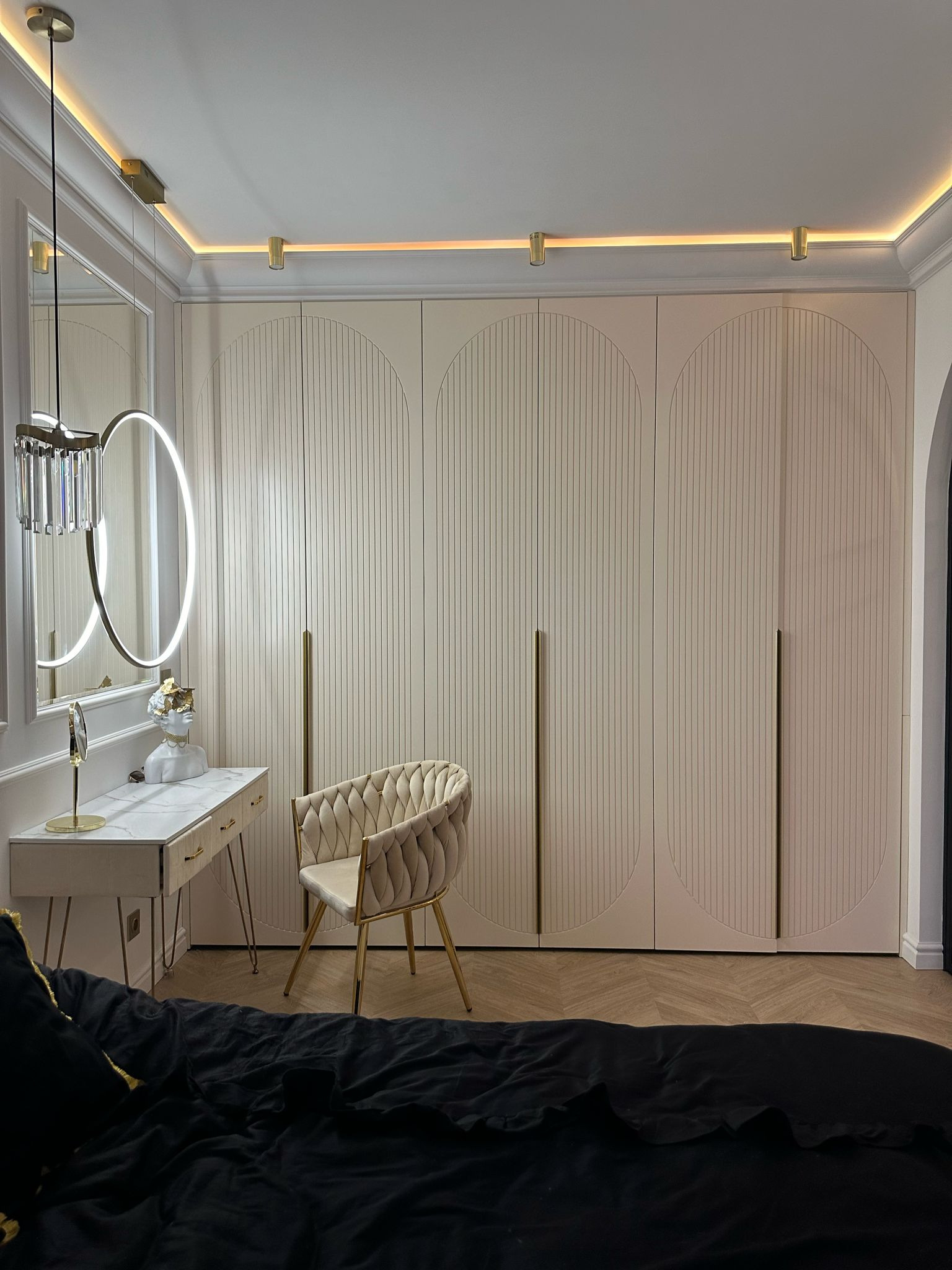 Elegancka sypialnia z beżową szafą wnękową z frezowanymi drzwiami, toaletką z marmurowym blatem i złotymi nogami oraz krzesłem w stylu glamour.