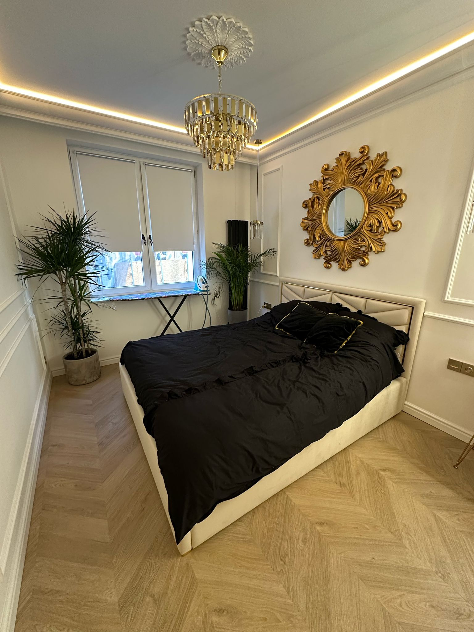 Elegancka sypialnia z łóżkiem w beżowej ramie, czarną pościelą, złotym lustrem nad wezgłowiem i kryształowym żyrandolem. Parkiet w jodełkę i rośliny doniczkowe.