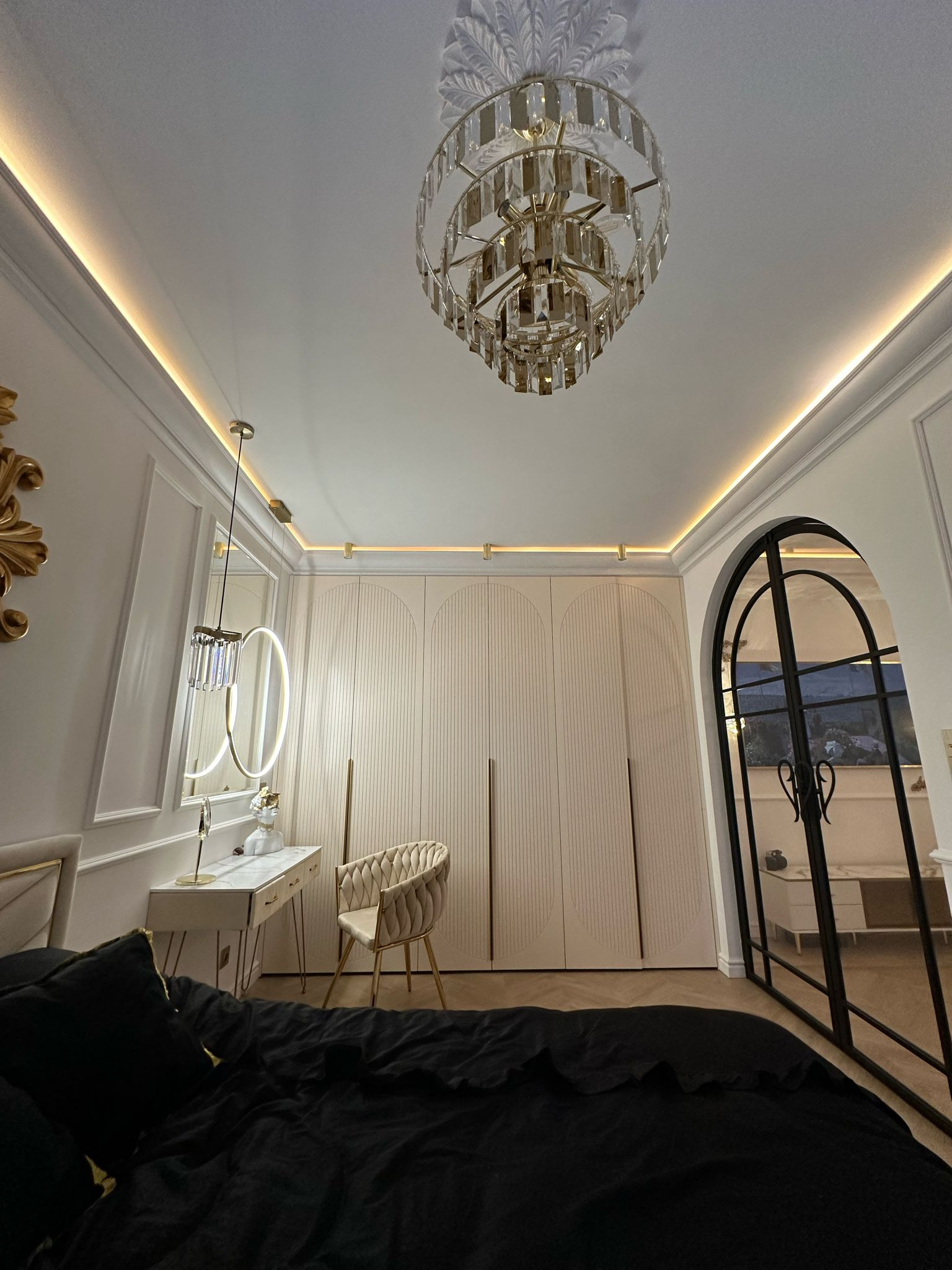 Elegancka sypialnia z garderobą w zabudowie, toaletką z okrągłym lustrem i kryształowym żyrandolem. Uwagę zwraca czarna pościel i drzwi łukowe.
