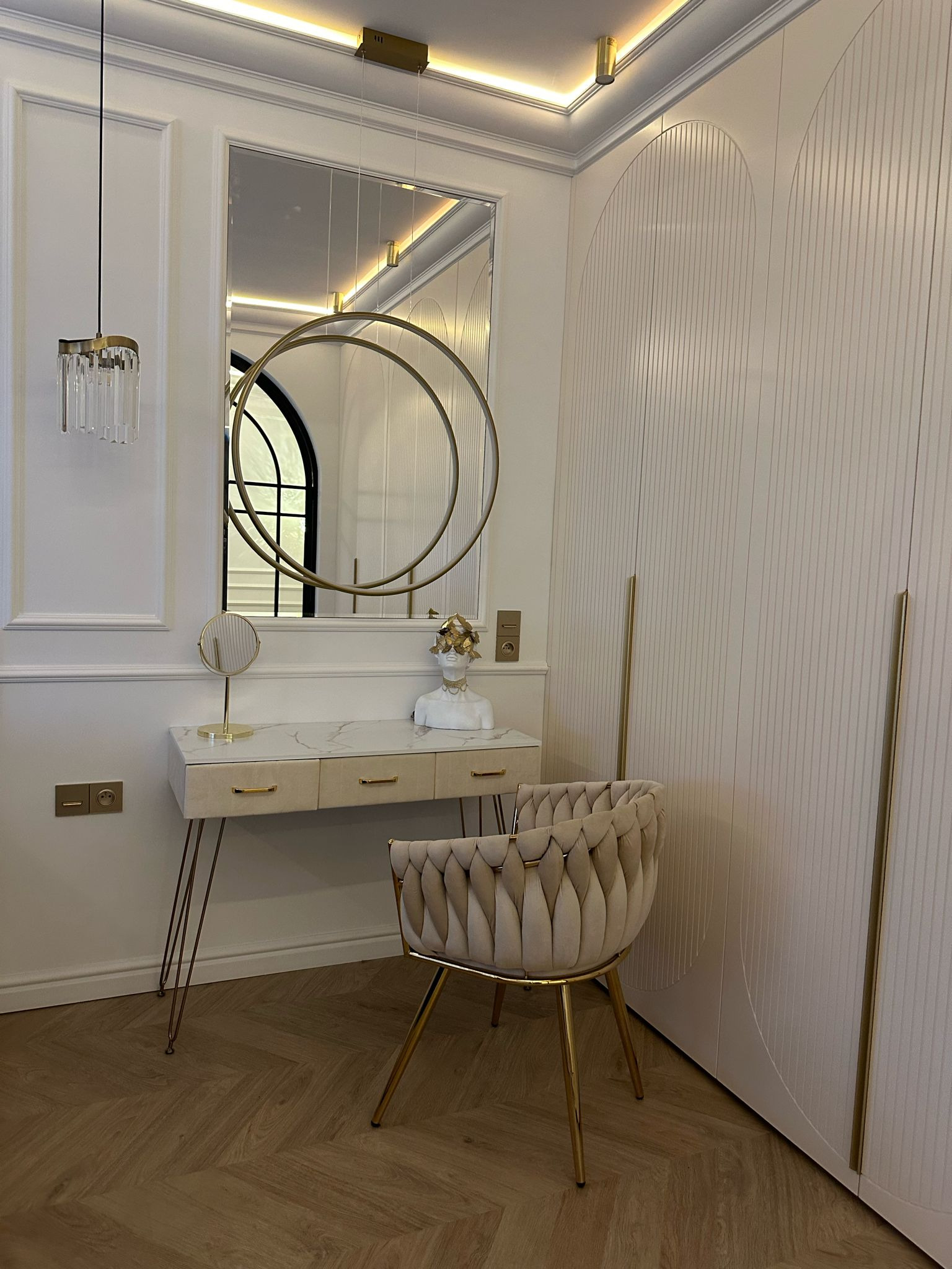 Elegancka toaletka z lustrem w złotej ramie i beżowym krzesłem na tle białej szafy z frezowanymi drzwiami. Całość w jasnej, stonowanej kolorystyce.