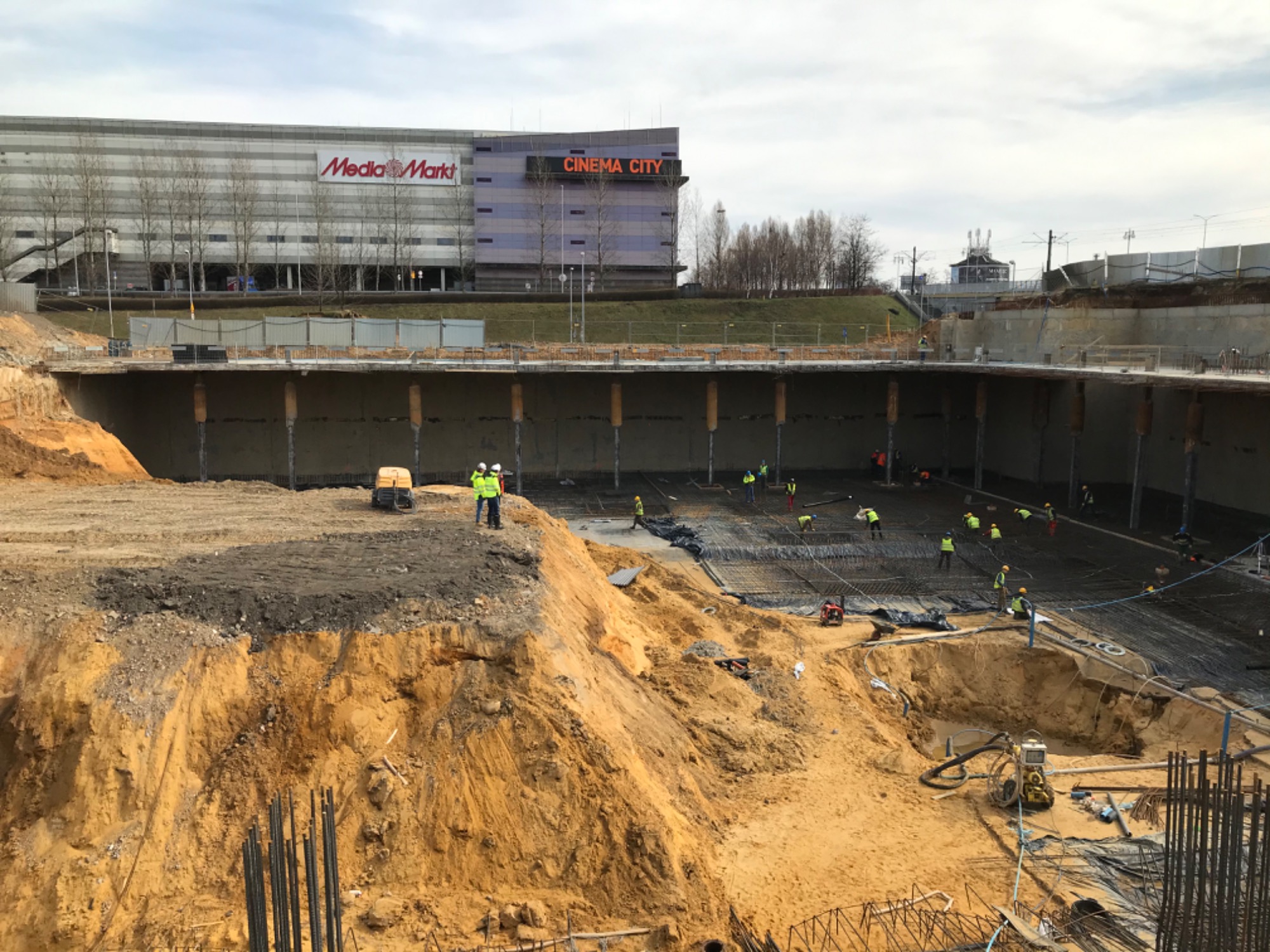 Budowa fundamentów pod budynek komercyjny, widoczni pracownicy w żółtych kamizelkach, zbrojenie i wykop z ziemią, w tle budynek Media Markt i Cinema City.