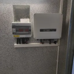 Biała skrzynka rozdzielcza PV AC/DC z bezpiecznikami i inwerter fotowoltaiczny zamontowane na szarej, chropowatej ścianie wewnątrz budynku. Widoczne przewody elektryczne i rurki osłonowe.