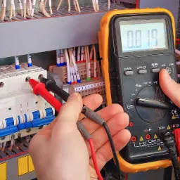 Miernik w dłoniach elektryka podczas pomiaru napięcia w rozdzielnicy; widoczne bezpieczniki, przewody i listwy zaciskowe