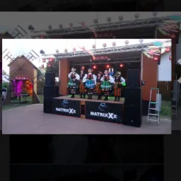 Zespół folklorystyczny w tradycyjnych strojach na scenie z nagłośnieniem i oświetleniem podczas występu plenerowego, obok dekoracyjna makieta wiatraka.