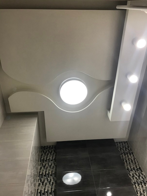 Nowoczesna łazienka z podwieszanym sufitem, okrągłym oświetleniem centralnym i listwą z punktowymi lampami. Na ścianach szare płytki i dekoracyjny pas mozaiki w odcieniach szarości i czerni.