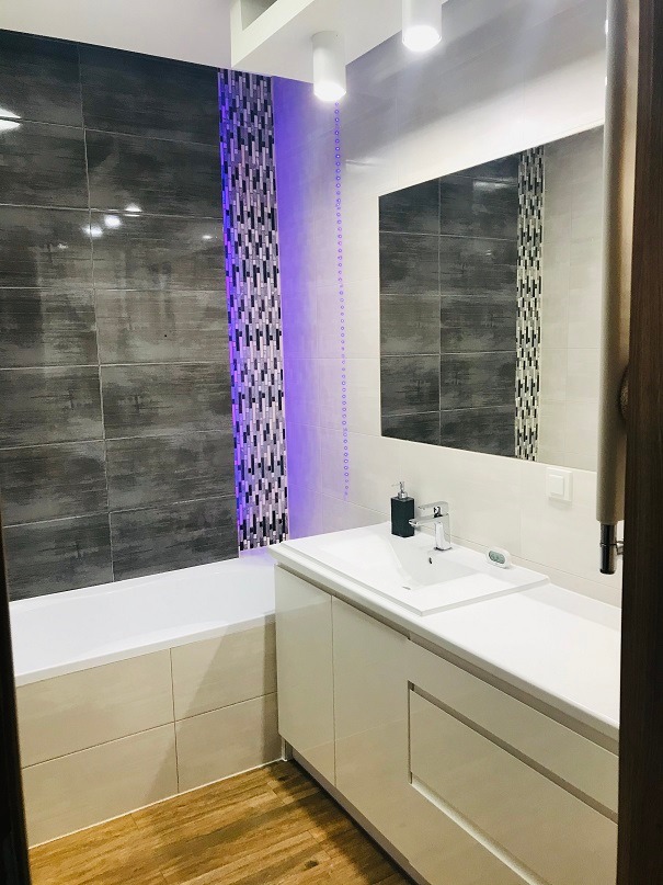 Nowoczesna łazienka z szarymi płytkami imitującymi beton, pionowym pasem mozaiki z podświetleniem LED, białą umywalką z szafką i drewnopodobną podłogą.