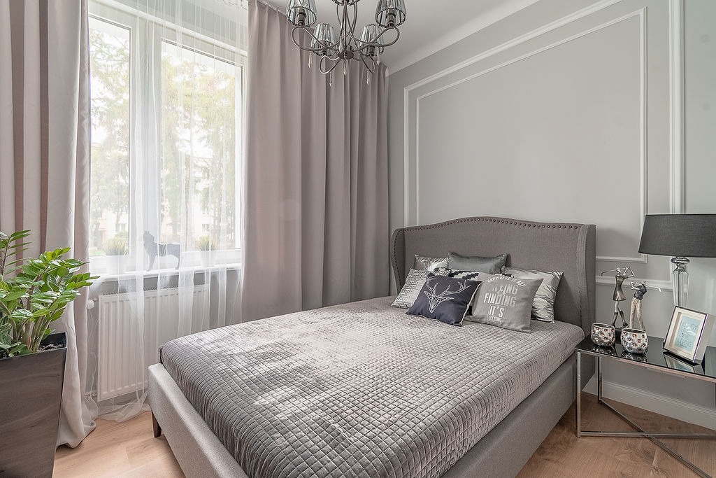 Elegancka sypialnia w odcieniach szarości z łóżkiem z pikowanym zagłówkiem, dekoracyjnymi poduszkami i rośliną doniczkową przy oknie.