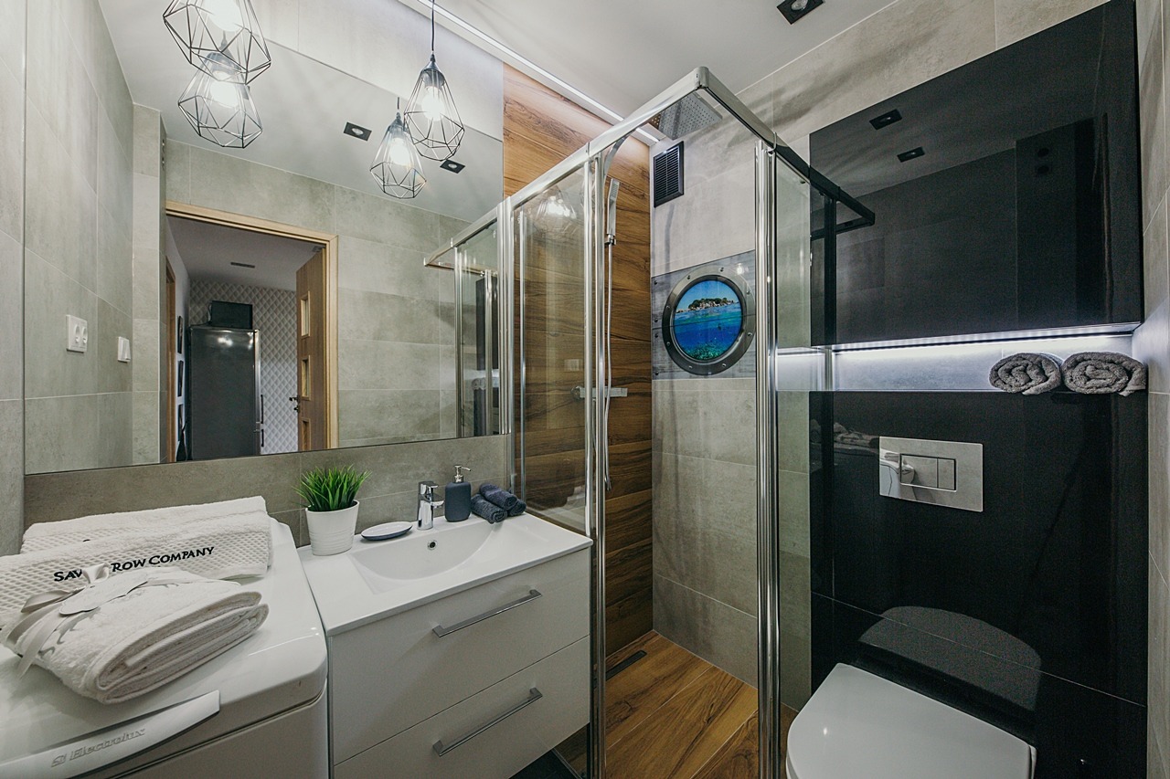 Nowoczesna łazienka z kabiną prysznicową, umywalką z szafką i lustrem, oświetlona wiszącymi lampami. Kabina z motywem okna statku z widokiem na morze. Na pralce leżą białe ręczniki.