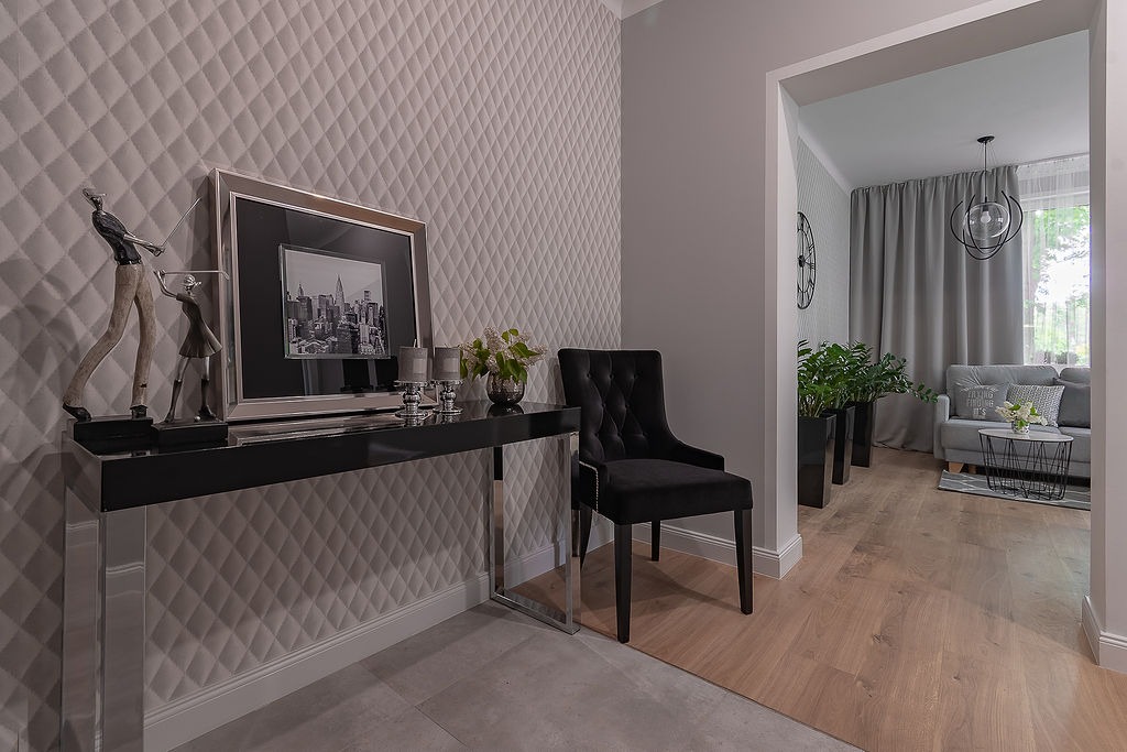 Elegancki przedpokój z pikowaną tapetą, czarnym stolikiem konsolowym z dekoracjami, czarnym krzesłem i widokiem na salon w odcieniach szarości.