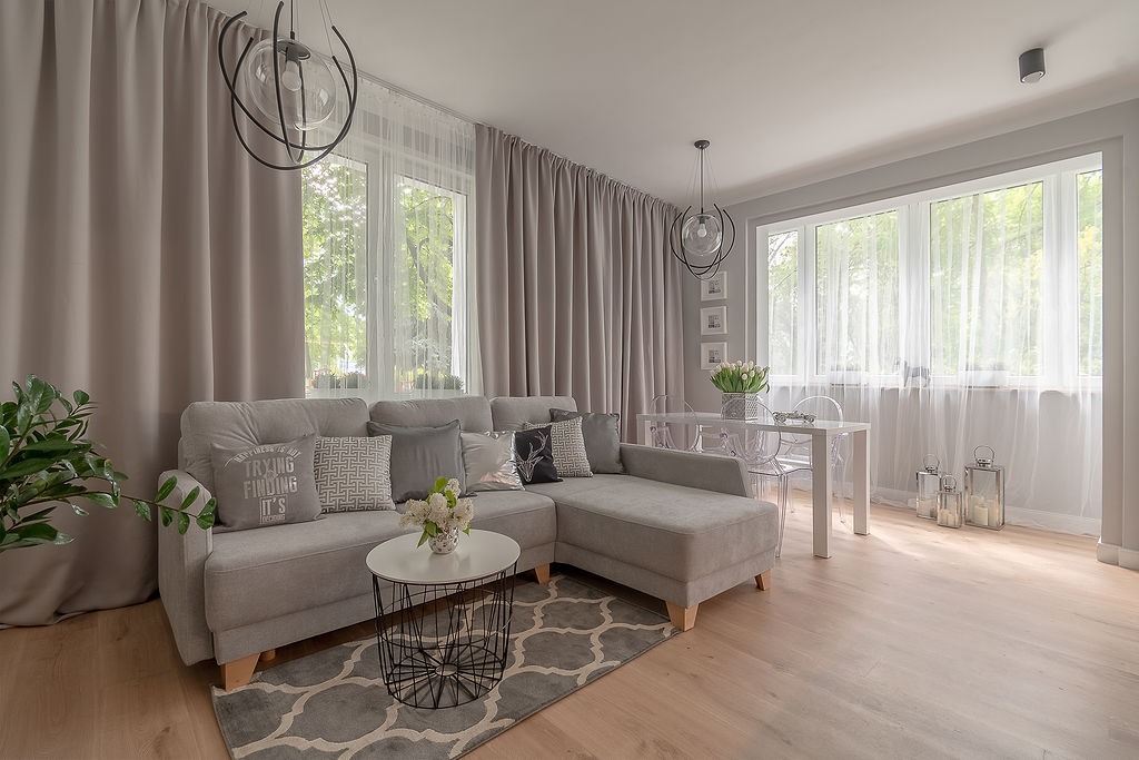 Jasnoszary salon z narożną sofą, okrągłym stolikiem kawowym na wzorzystym dywanie, białym stołem z przezroczystymi krzesłami i długimi, beżowymi zasłonami przy oknach.