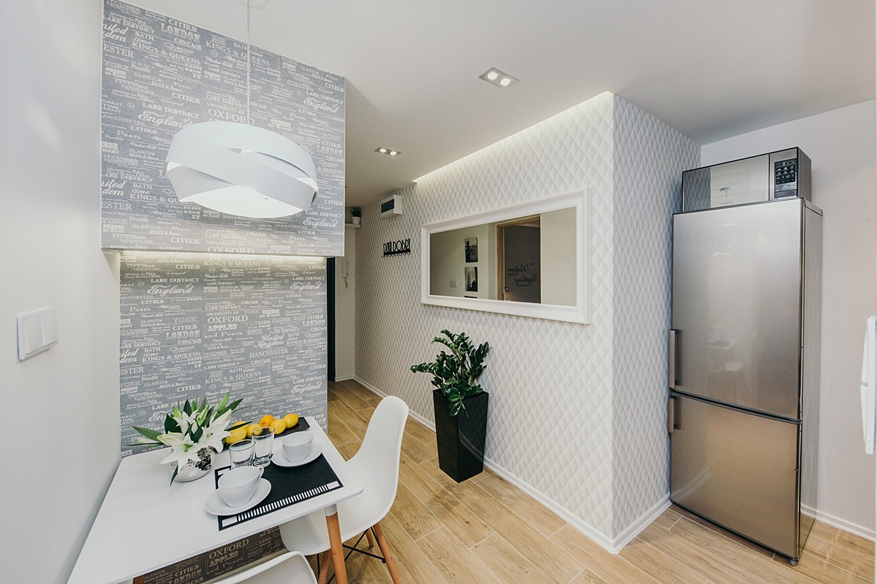 Jasne wnętrze mieszkania z tapetą w angielskie motywy, minimalistycznym stołem z kwiatami i owocami, oraz srebrną lodówką i dekoracyjną ścianą z lustrem.