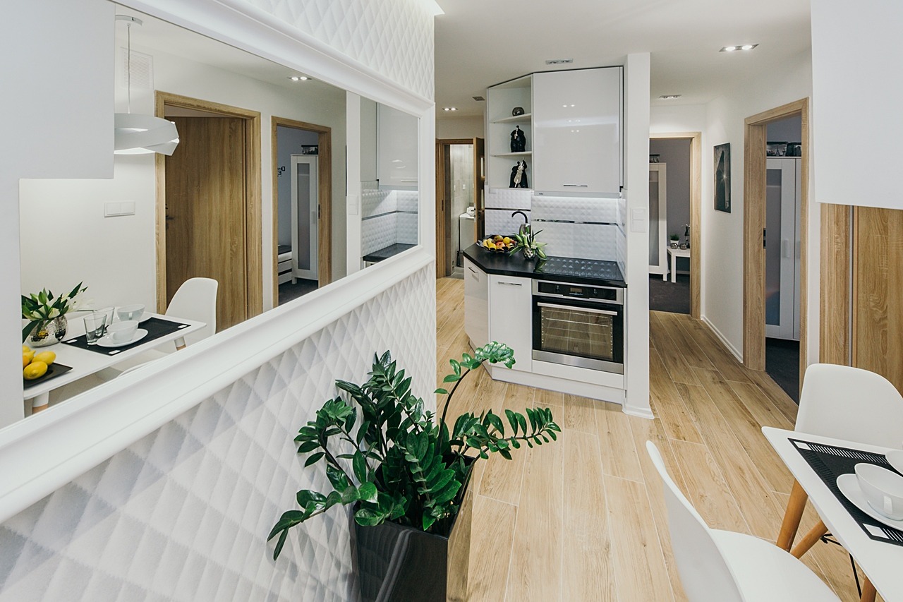 Nowoczesne wnętrze mieszkania z otwartą kuchnią, widoczne białe szafki kuchenne z czarnym blatem, drewnopodobne panele na podłodze, biała ściana z geometrycznym wzorem, duże lustro i roślina...