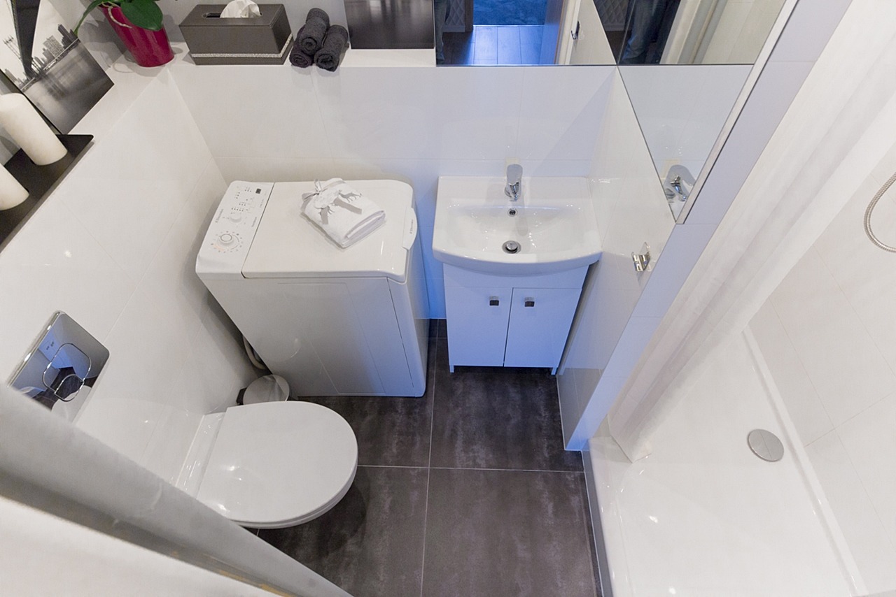 Mała, ale funkcjonalna łazienka w odcieniach bieli i szarości z pralką ładowaną od góry, umywalką z szafką, toaletą i kabiną prysznicową, wykończona płytkami podłogowymi w kolorze grafitowym.