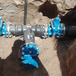Złożony system rur z niebieskimi zaworami i srebrnymi śrubami, umieszczony w wykopie z ziemi.