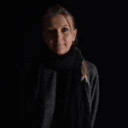 Portret kobiety z blond włosami spiętymi w kucyk, ubrana w szary sweter w kratę i czarny szal, oświetlona z góry w ciemnym otoczeniu.