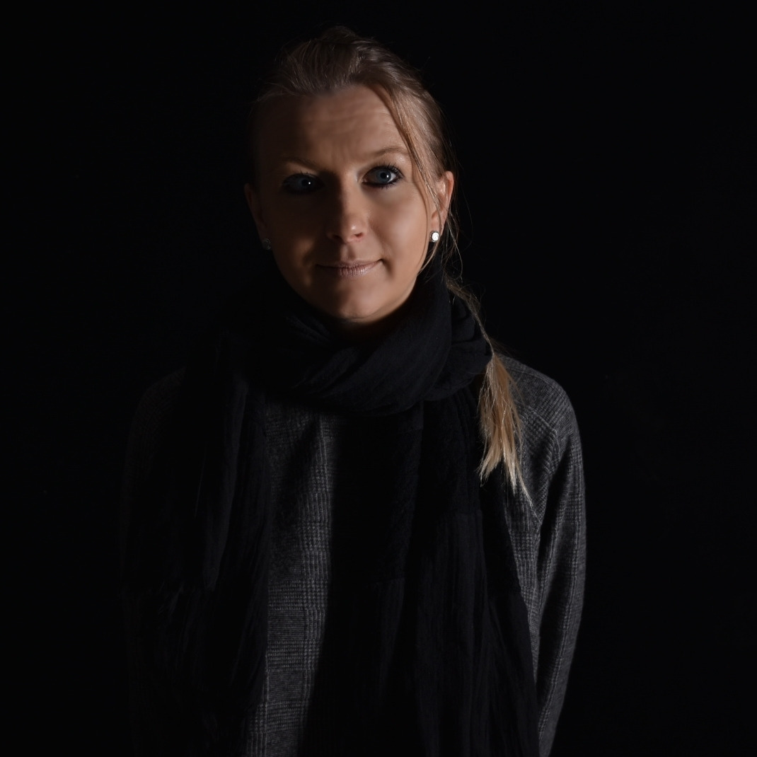 Portret kobiety z blond włosami spiętymi w kucyk, ubrana w szary sweter w kratę i czarny szal, oświetlona z góry w ciemnym otoczeniu.